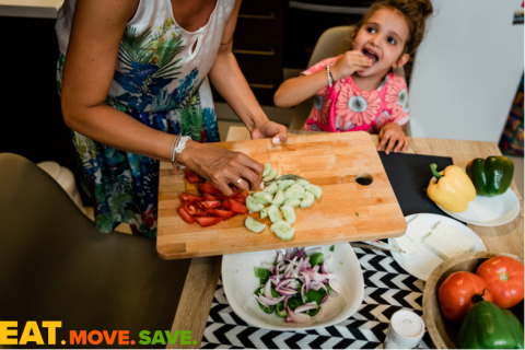 A child sits a kitchen table eating a sliced piece of vegetable. A woman stands beside her and is pushing cut cucumbers and tomatoes from a cutting board to a bowl of salad on the table. 