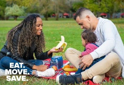 mom, dad and child sitting on a picnic blanket in the park. mom offers a banana to the child while smiling.
