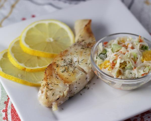 cooked fish with dill seasoning, side of coleslaw and lemon slices on white plate