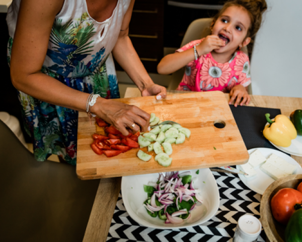 A child sits a kitchen table eating a sliced piece of vegetable. A woman stands beside her and is pushing cut cucumbers and tomatoes from a cutting board to a bowl of salad on the table. 