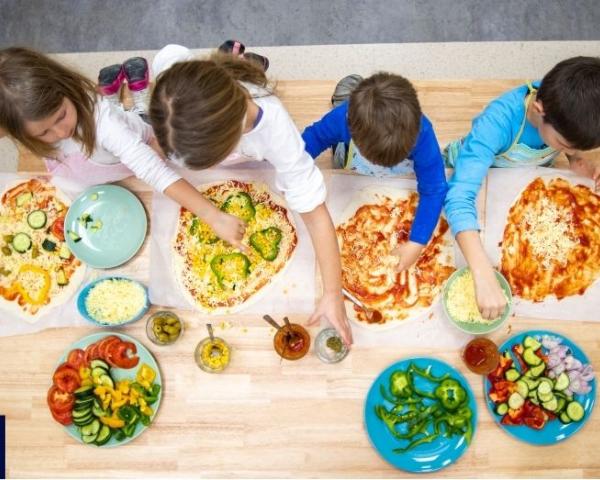 Four kids add different vegetable toppings to their own pizzas. 