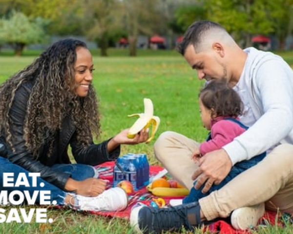 mom, dad and child sitting on a picnic blanket in the park. mom offers a banana to the child while smiling.