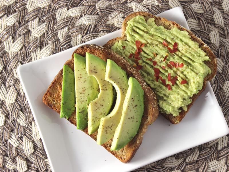 two slices of toast, one topped with sliced avocado and spices and one with smashed avocado with red pepper flakes