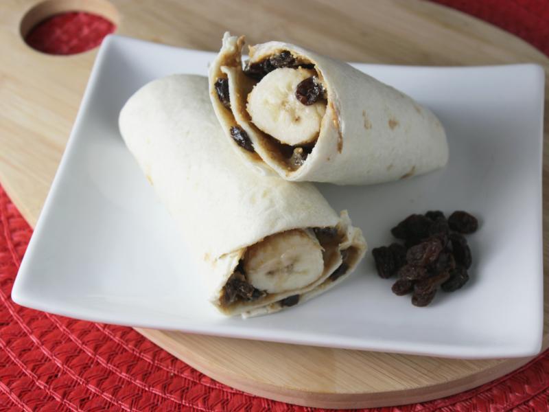 tortilla rolled with peanut butter, banana, and raisins cut in half and stacked on white plate