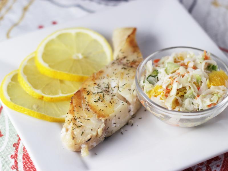 cooked fish with dill seasoning, side of coleslaw and lemon slices on white plate