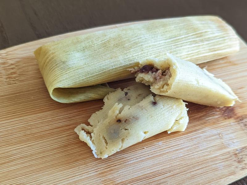 cooked tamale split in half with tamale wrapped in cornhusk in background