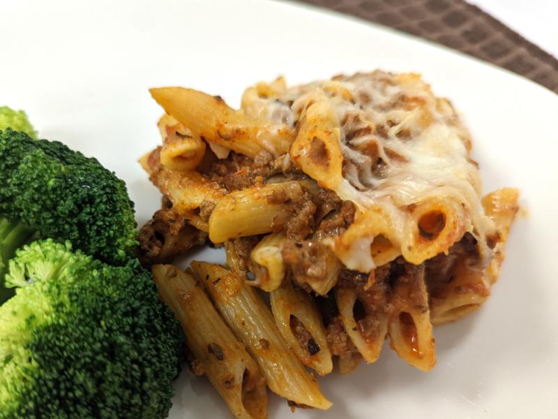 penne pasta mixed with tomato sauce and ground meat and topped with cheese on white plate with side of broccoli