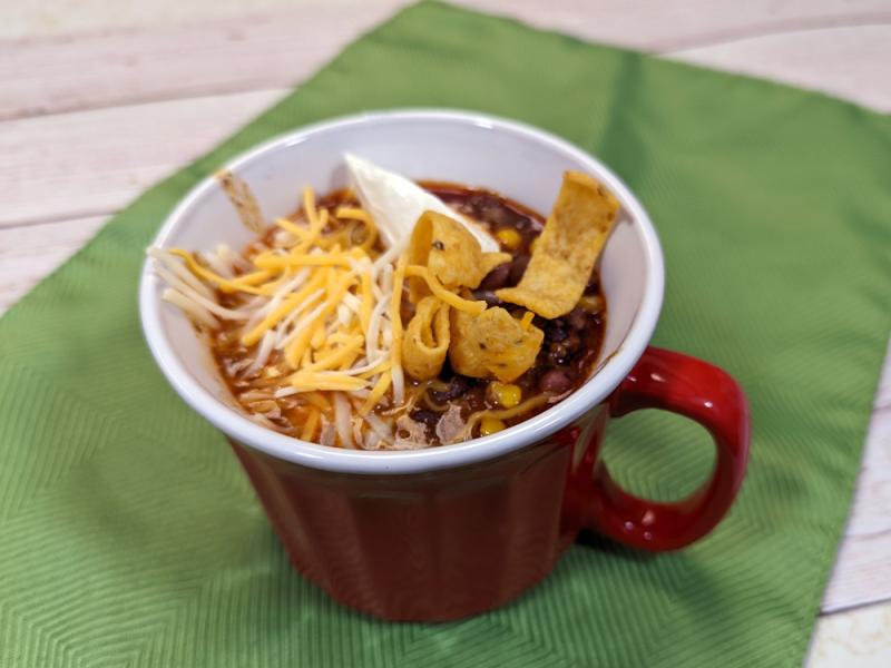red mug with taco soup, topped with cheddar cheese, corn chips, and sour cream, on a green napkin