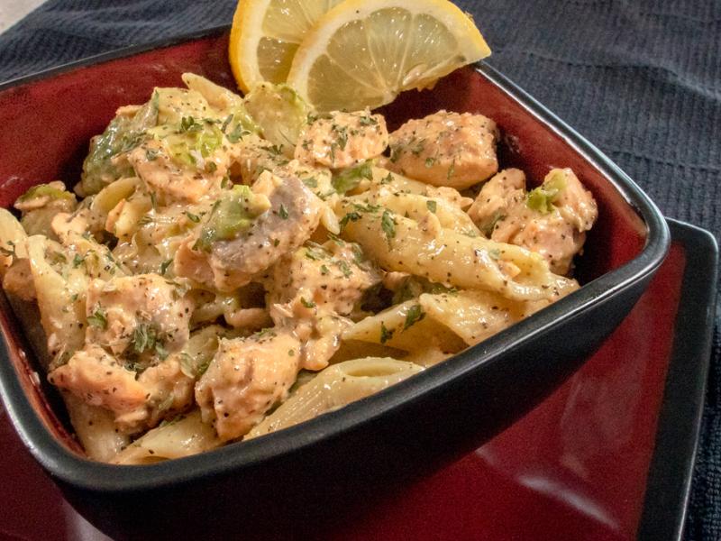salmon and pasta with creamy sauce in a dark red bowl with slices of lemon on the side