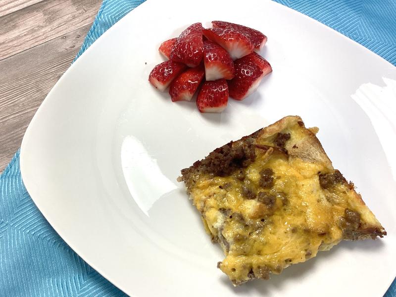 square of cooked breakfast casserole on white plate with a side of sliced strawberries