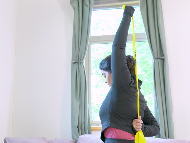 Person stretching an exercise band behind back with one hand overhead and other at base of back.