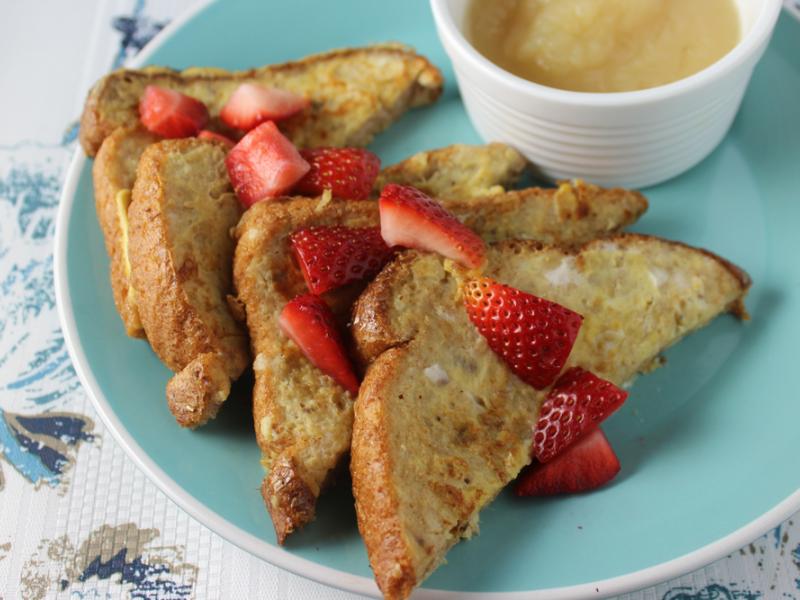 french toast topped with sliced fresh strawberries served with a cup of applesauce on a teal plate